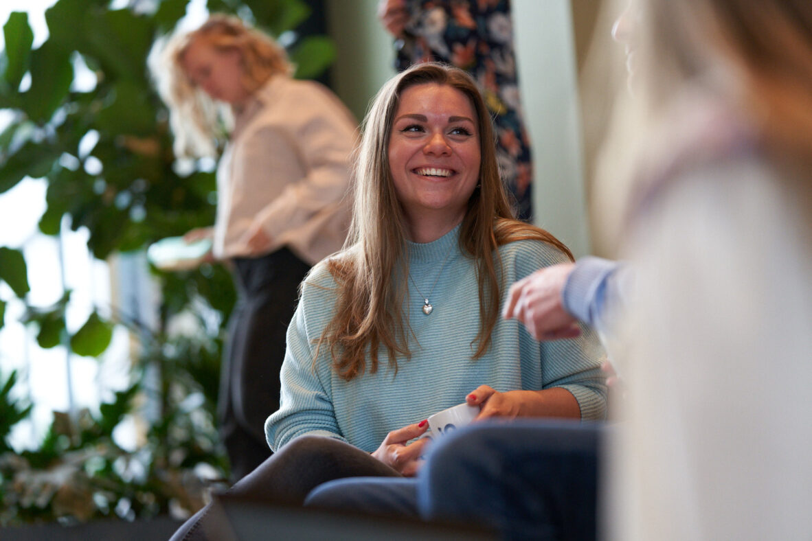 Een lachende vrouw met lang haar zit op een bank en houdt een mok vast, met een andere persoon op de achtergrond.