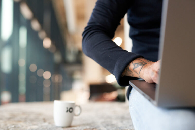 Een persoon met een tatoeage typt op een laptop, met een kleine koffiepot op tafel in een lichte omgeving.