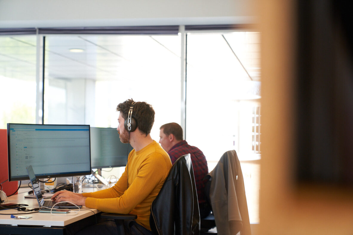 Een man met een geel shirt en hoofdtelefoon werkt aan een computer in een moderne kantooromgeving.