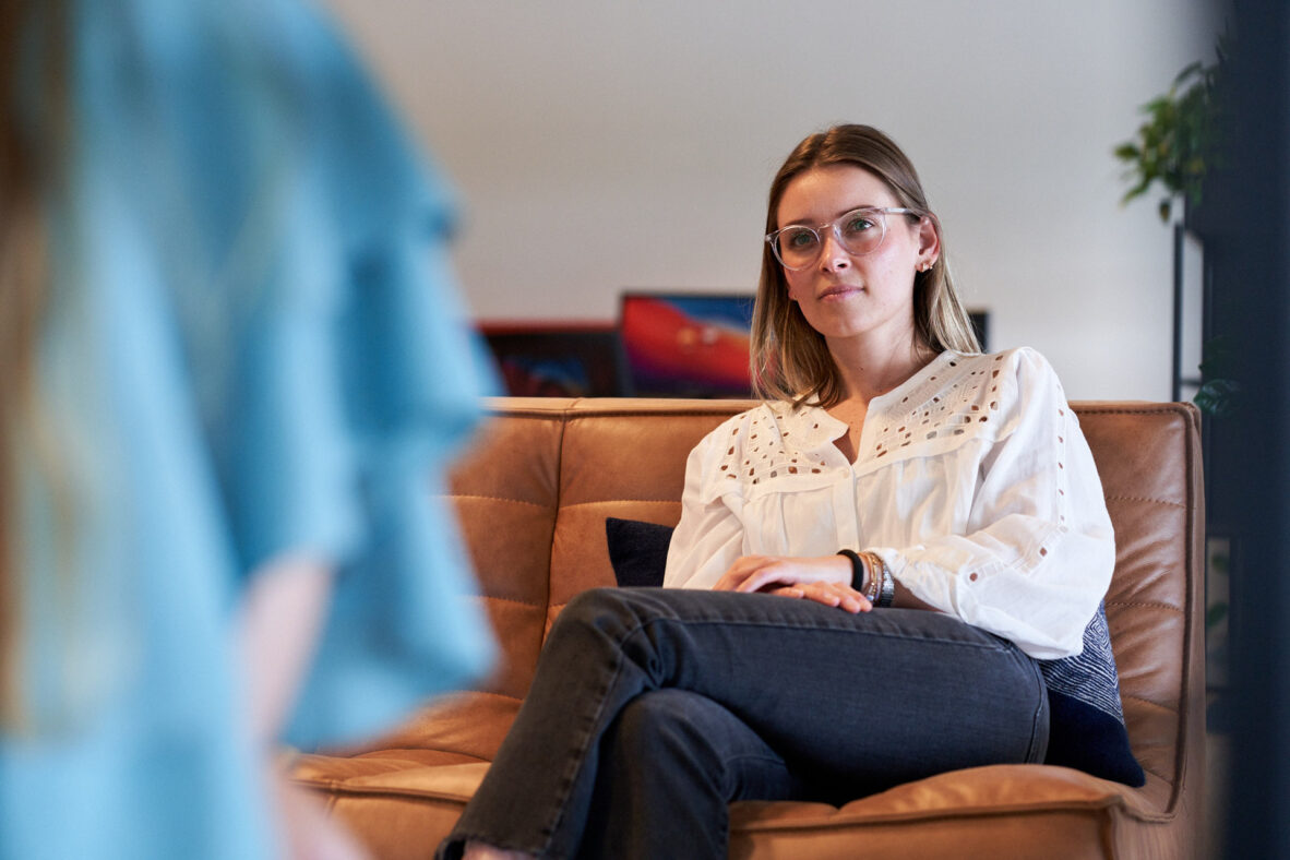 Een vrouw in een wit blouse zit op een bank, kijkend naar een andere persoon buiten beeld.