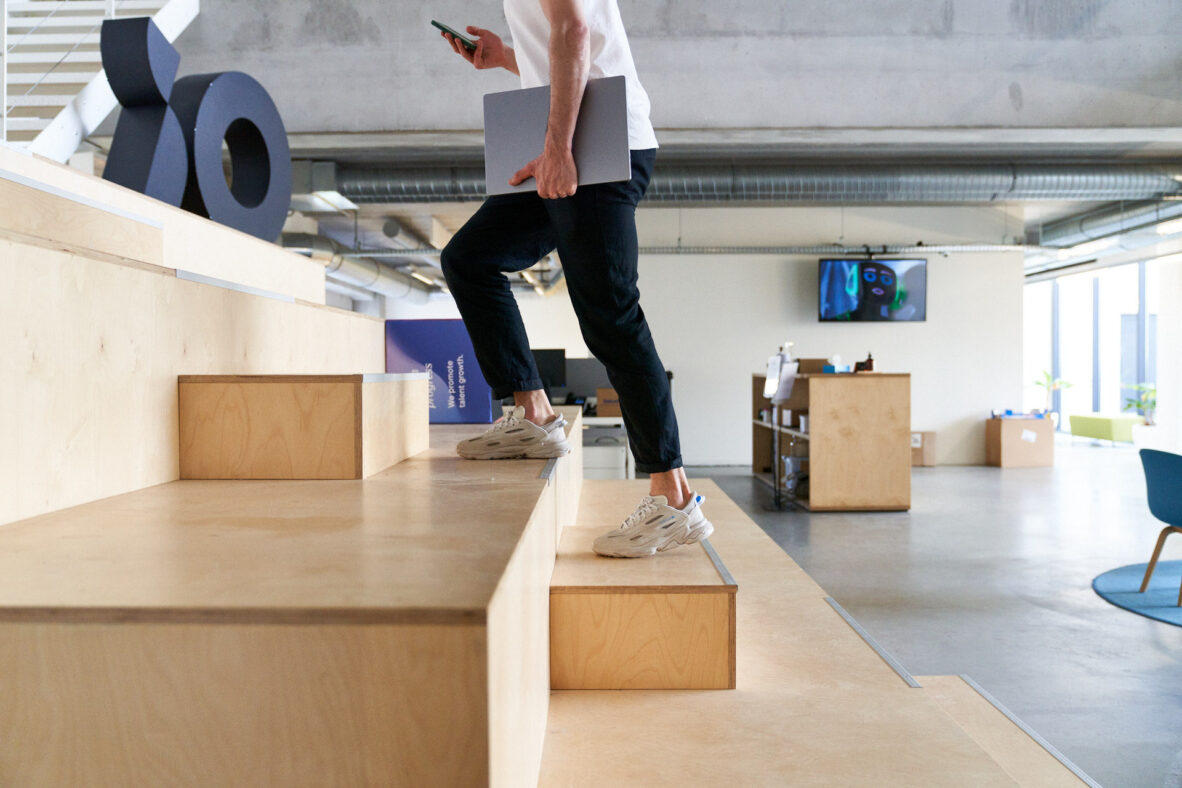 Een persoon loopt een houten trap op in een modern kantoor met een laptop en een telefoon in de hand.