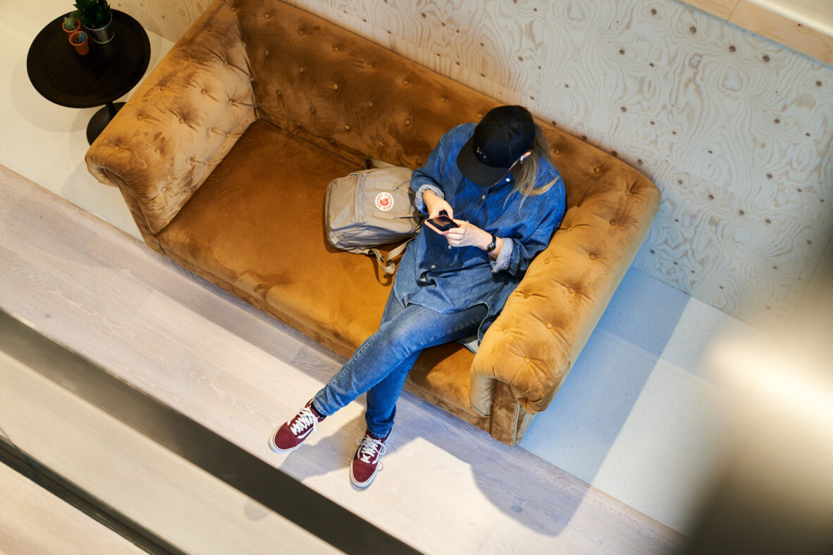 Vrouw in blauw denim outfit en pet zit op een bruine bank, kijkend naar haar telefoon met een rugzak naast zich.