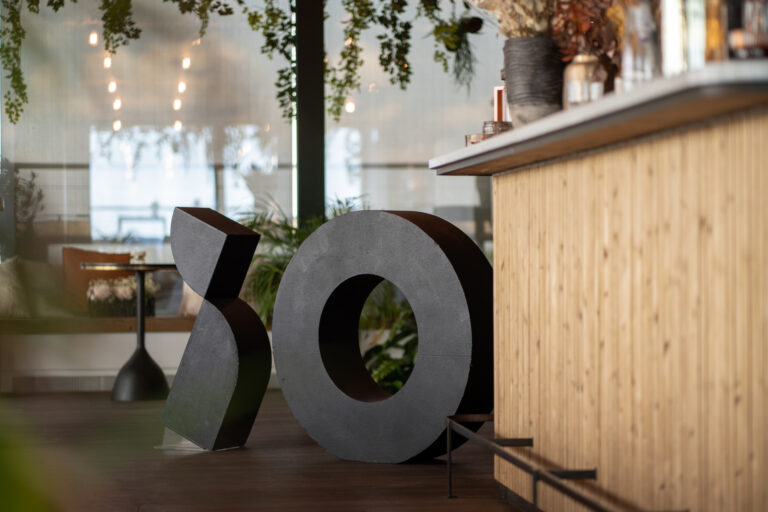 Een zwart sculptuur in de vorm van "00" staat voor een houten bar in een moderne ruimte met planten.