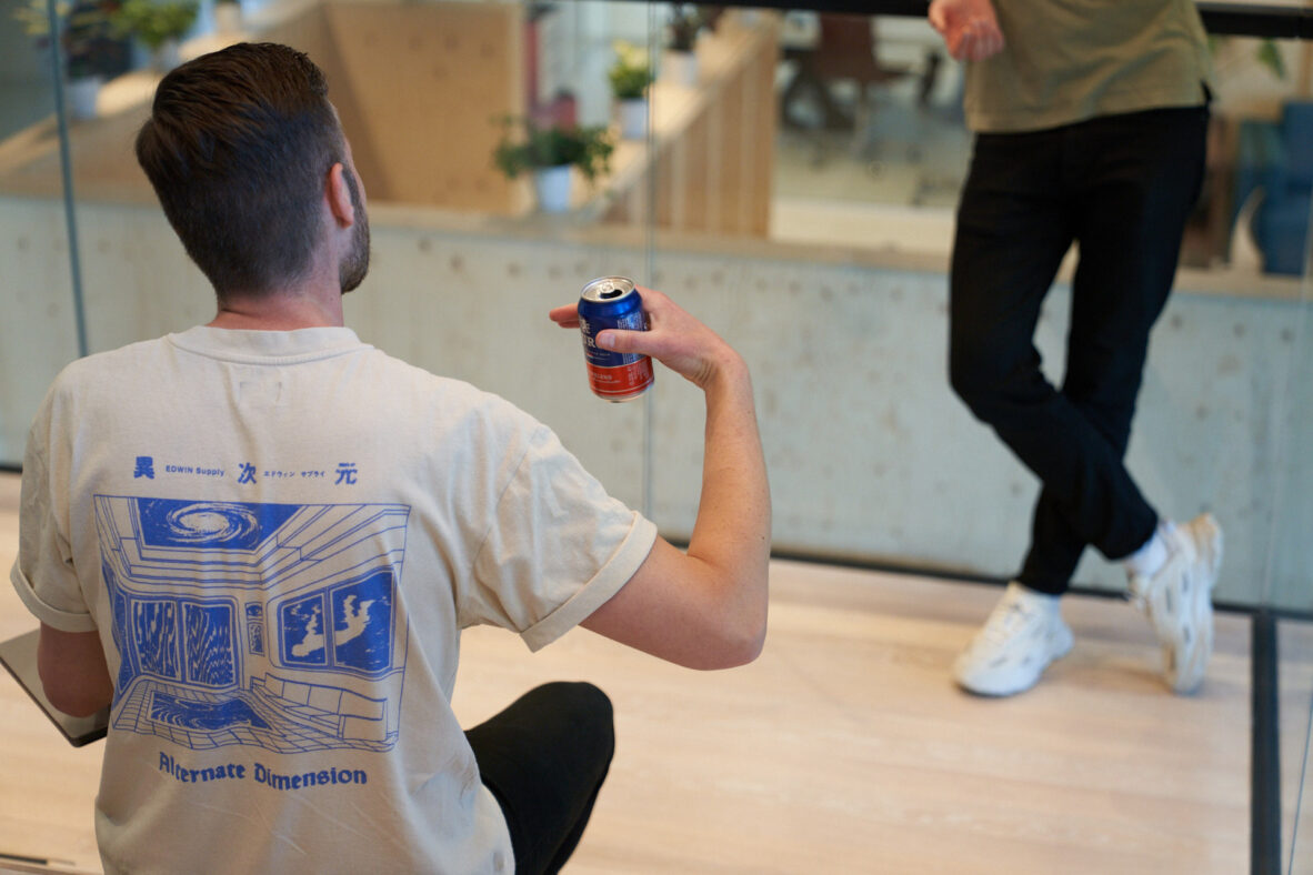 Een man in een beige T-shirt met een kunstafbeelding, terwijl hij een blikje drank vasthoudt en naar een ander kijkt.