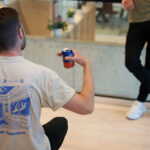 Een man in een beige T-shirt met een kunstafbeelding, terwijl hij een blikje drank vasthoudt en naar een ander kijkt.